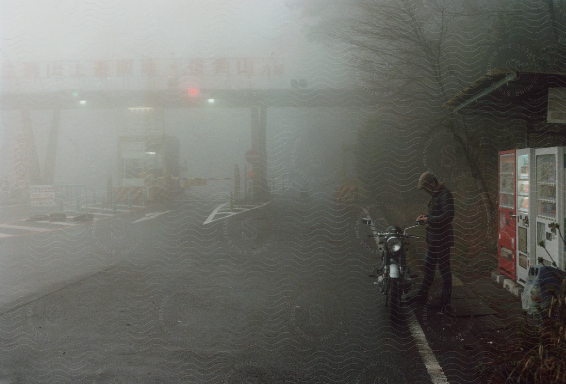 A man is standing near his motorcycle and vending machines along the side of a foggy road with a toll booth