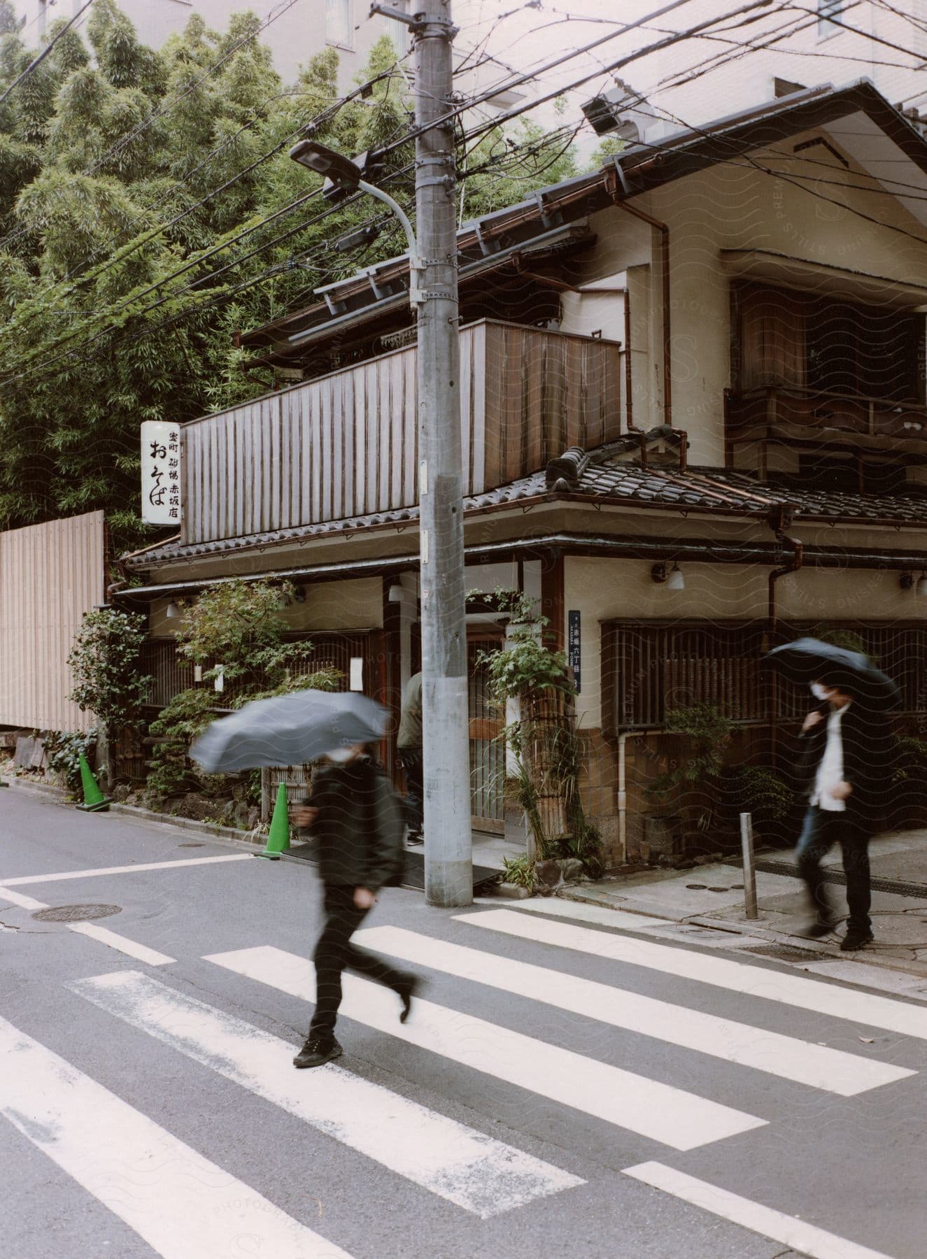 Two pedestrians with masks cross a street carrying black umbrellas