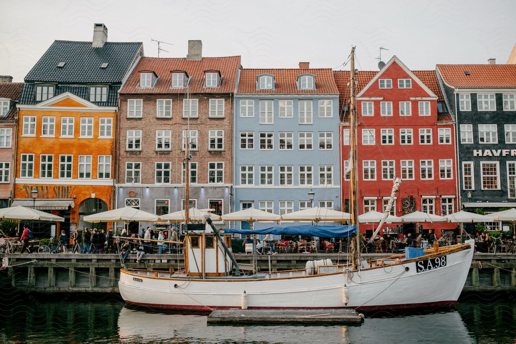 A boat docked in front of a row of colorful Nyhavn Harbor buildings, people strolling under umbrellas and sitting at outdoor restaurants.