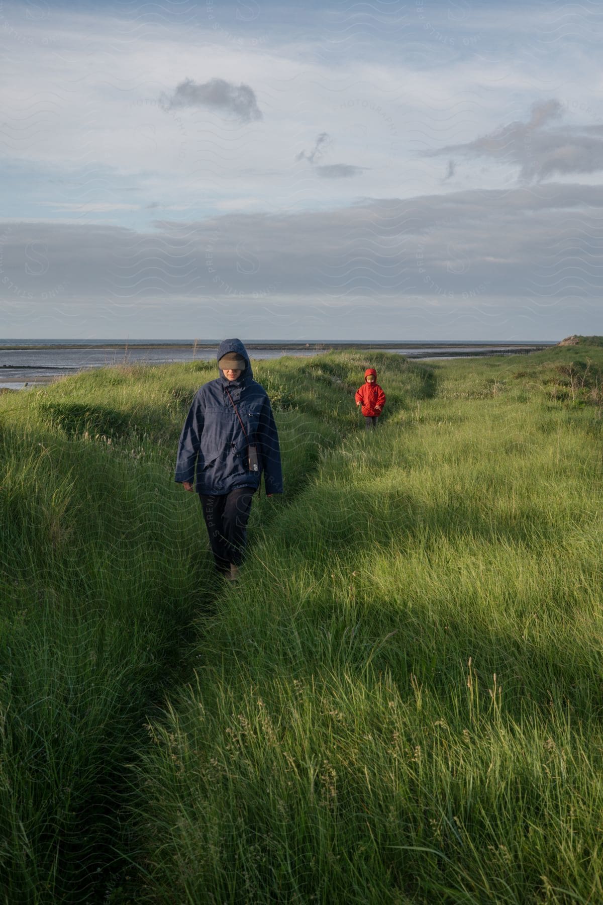 A woman and child walking on a thin trail in tall green grass away from the ocean