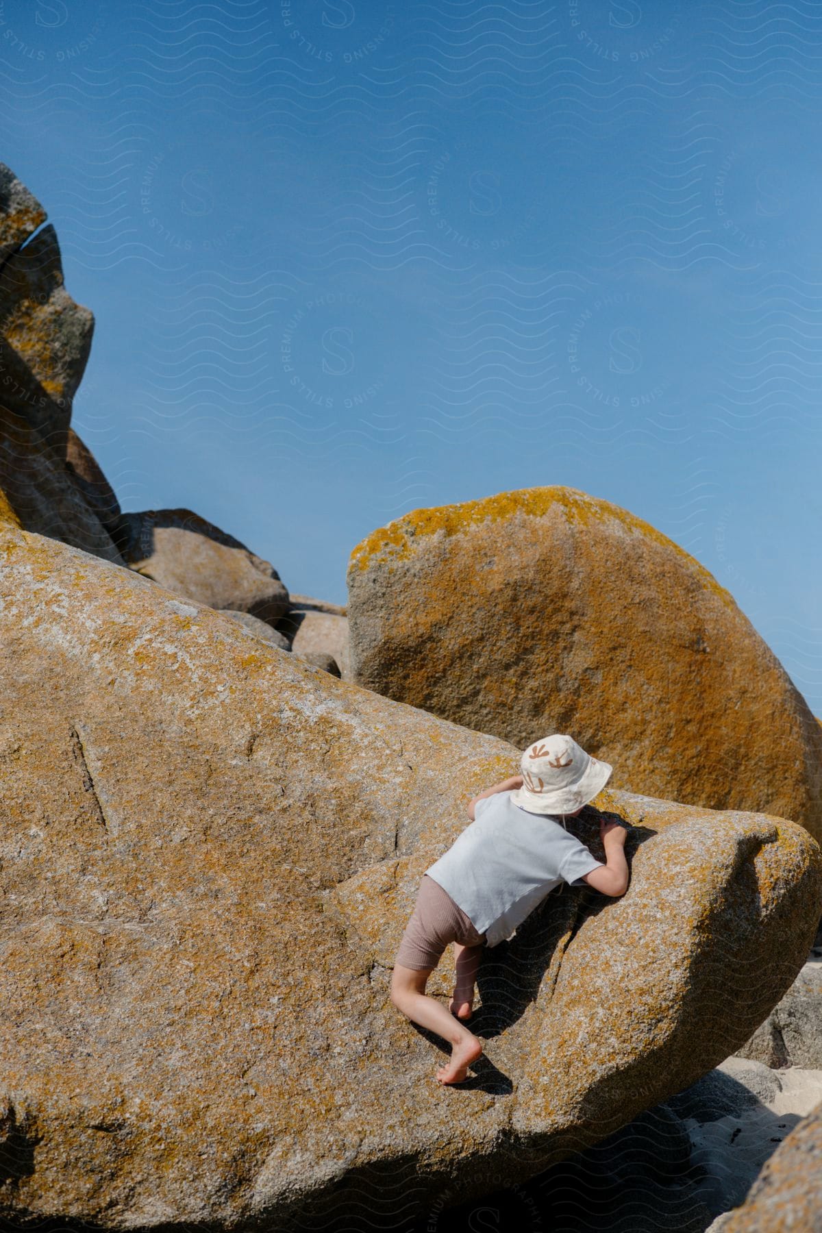 A barefoot child in shorts and bucket hat climbing rocks on a sunny day
