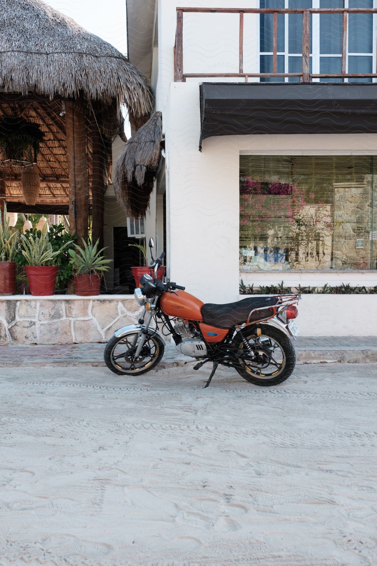 An orange accented motorcyle parked on a sandy dirt street in front of a building and thatch roof hut