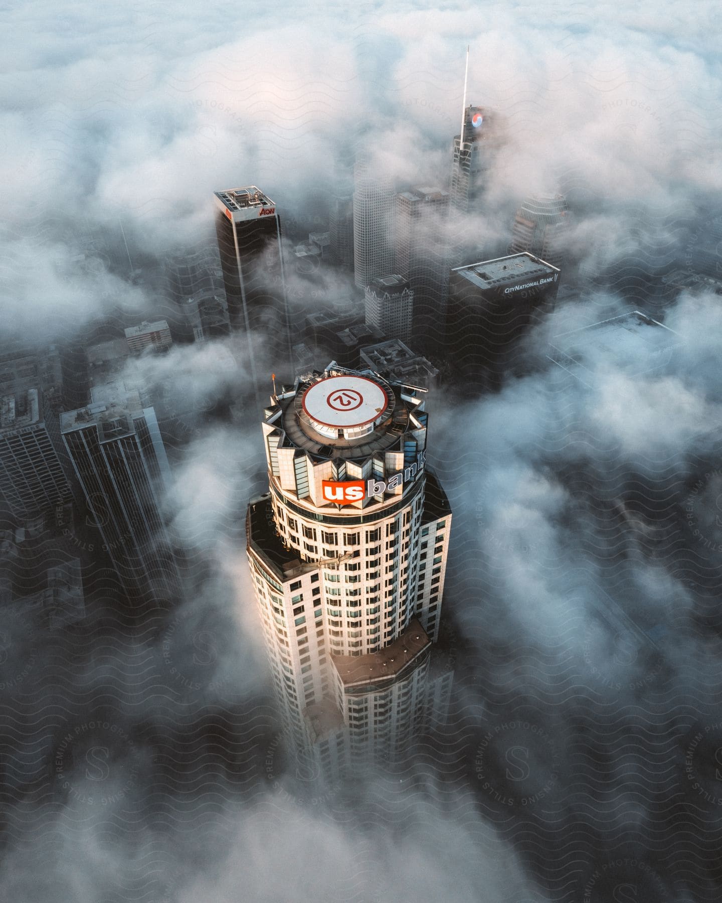 On a sunny day, the top of the US Bank skyscraper stands tall, with "12" clearly marked on the helipad. The building is surrounded by other towering skyscrapers, and the atmosphere is veiled in a mystical fog