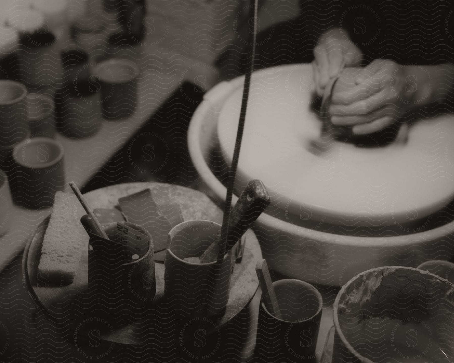 Pottery tools in cups and ceramic cups on a shelf near a person's hands working with clay on a pottery wheel