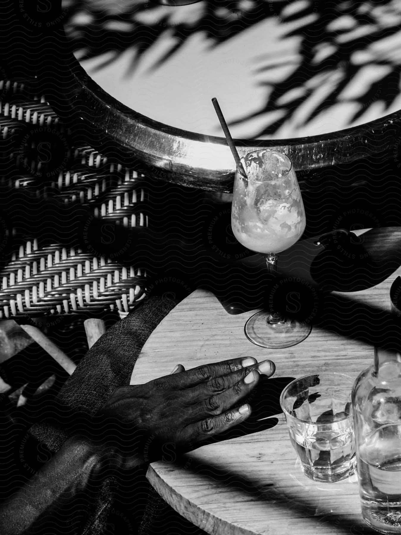 person sits at an outdoor cafe, their hand resting on the table, a glass of wine with a straw and cream in front of them, the shadows from a tree branch casted across the table