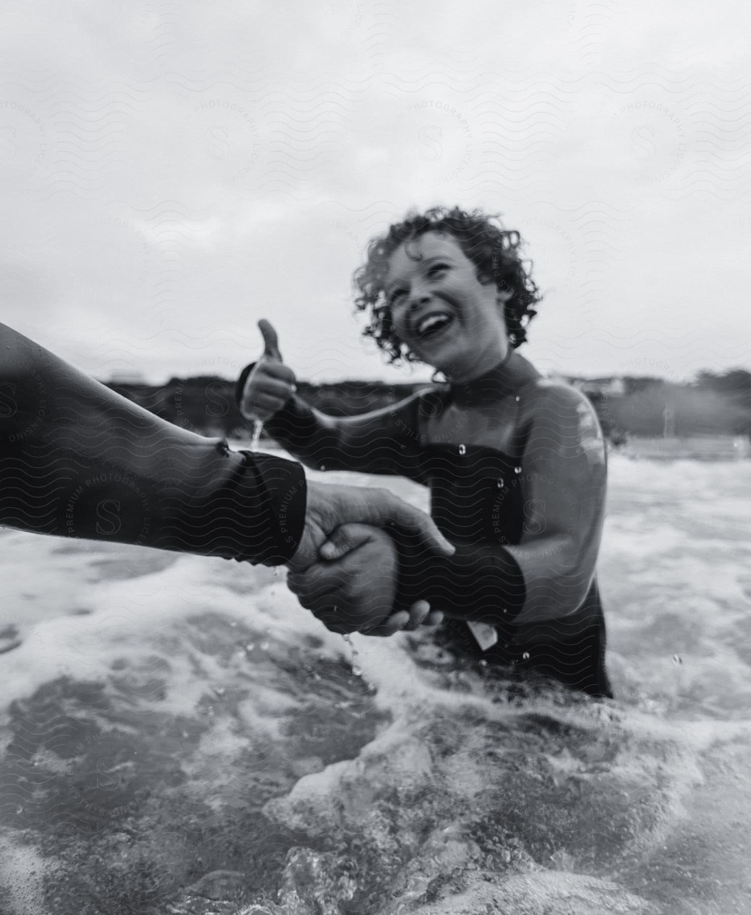 A Young Female With Curly Hair Is Standing In The Ocean While Smiling And Giving A Thumbs-up Gesture