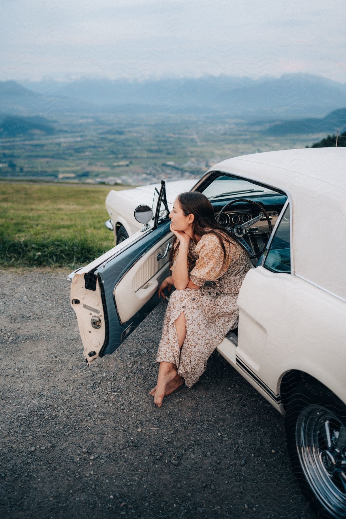 A woman in a sun dress sits in the driver's seat of her car on a hill, overlooking a vast expanse of farmland.