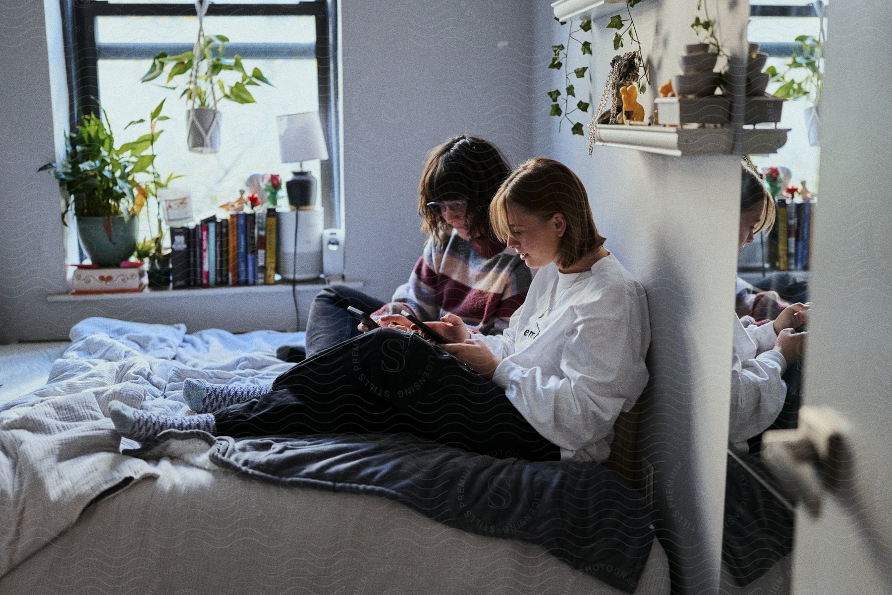 two teens look at their phones while sitting in bed together