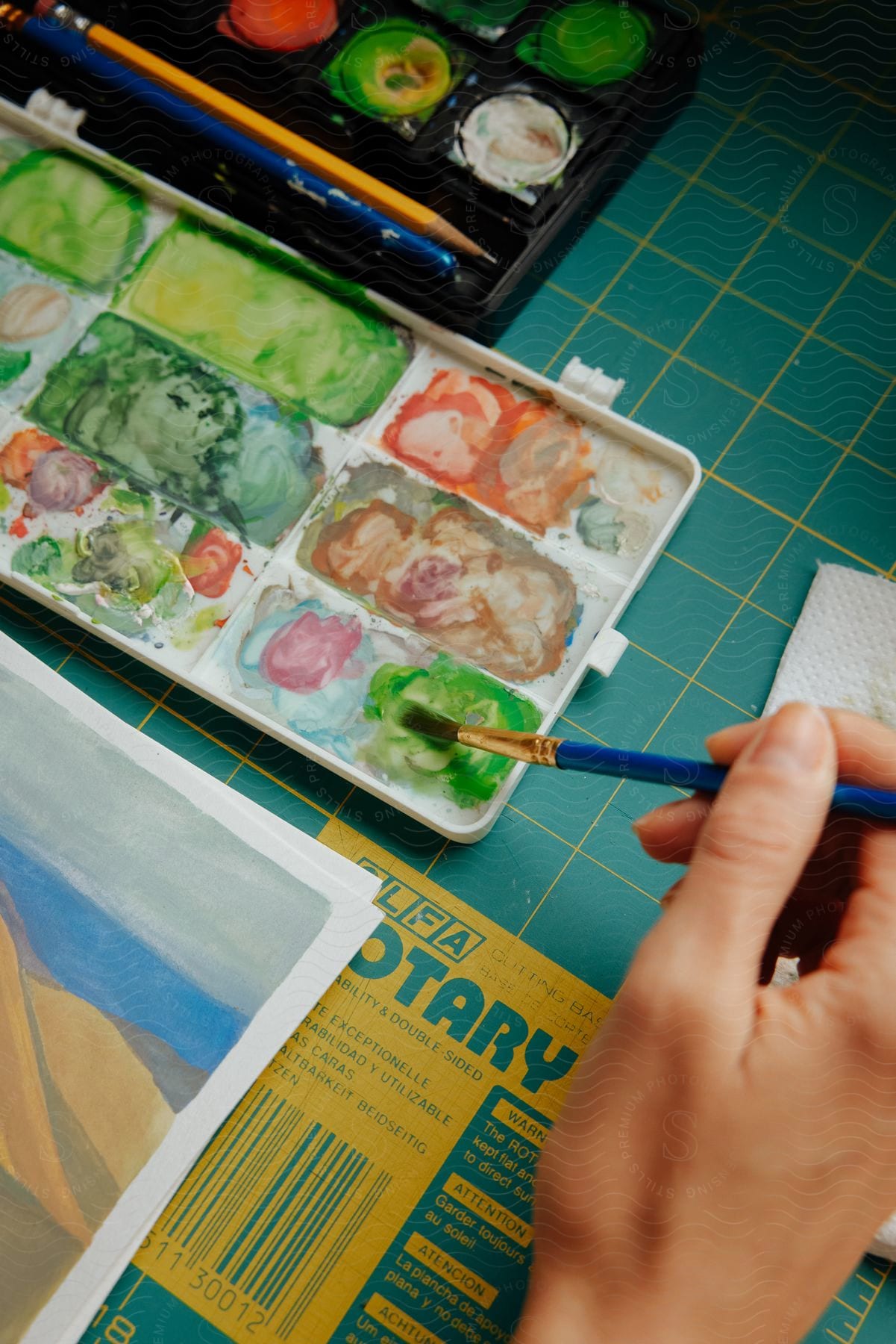 An artist sitting at a table is holding a paintbrush and mixing colors in a paint tray