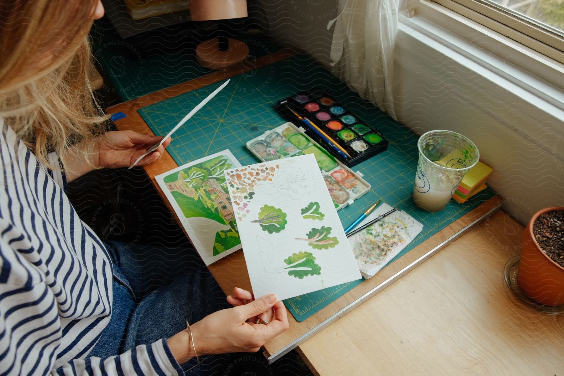 A woman in a long-sleeved striped shirt sits at a table near a window, looking at her artwork, a tray of paints of various colors and a paintbrush nearby.