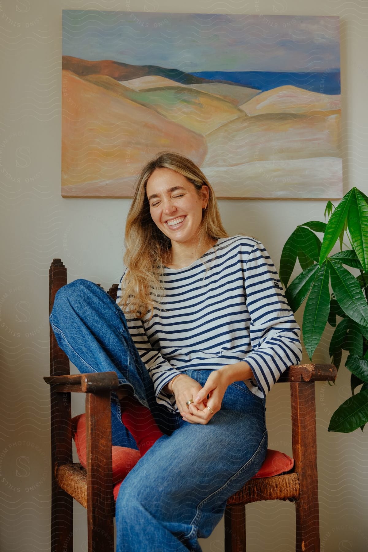 A young woman with long blonde hair is sitting in a chair next to a plant and a painting on the wall as she laughs