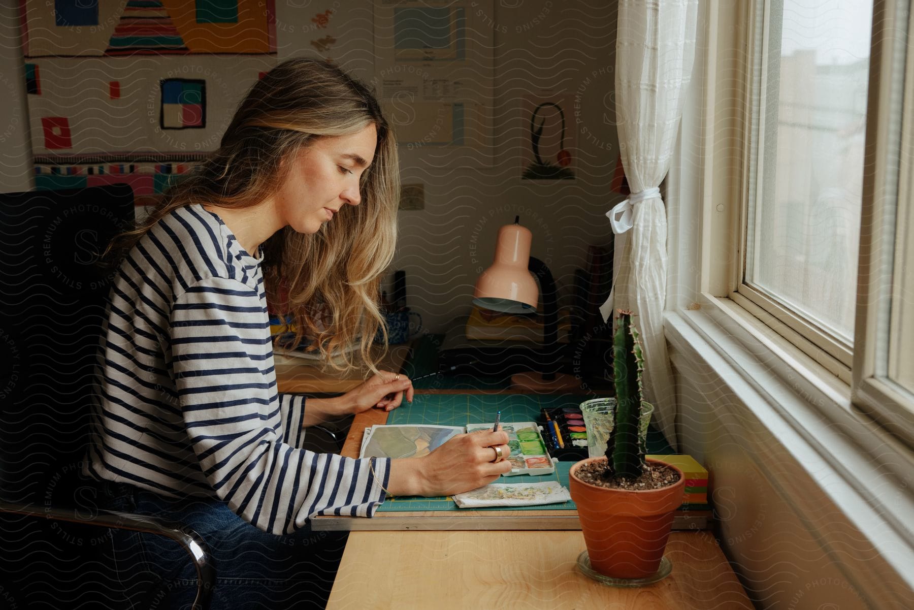 A woman with long blonde hair is sitting at a table in front of a window holding a paint brush and painting a picture