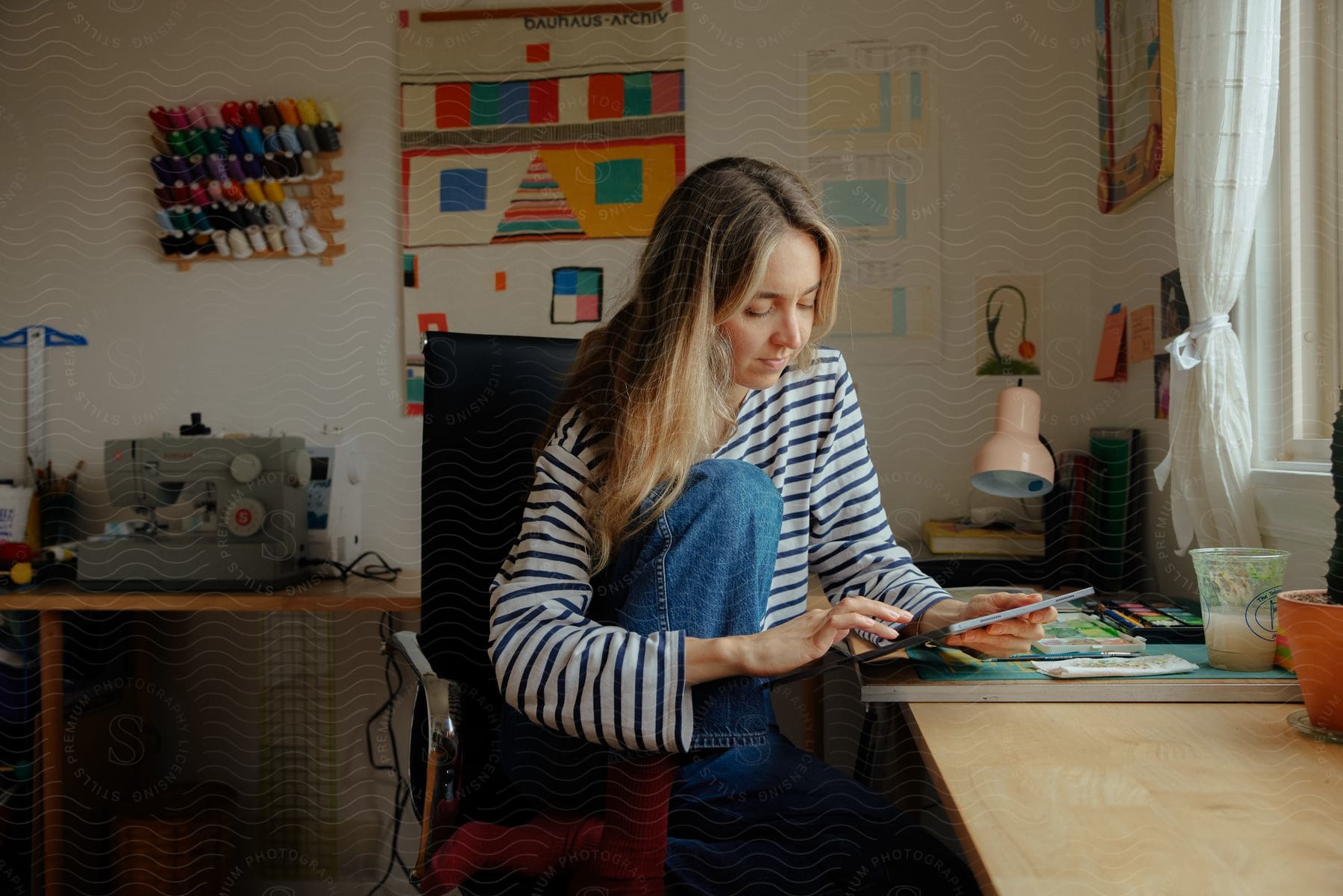 A woman in a striped shirt, sitting at a table near a window, looking at a tablet, with a tray of paints and a paintbrush in front of her, and a sewing machine in the background.