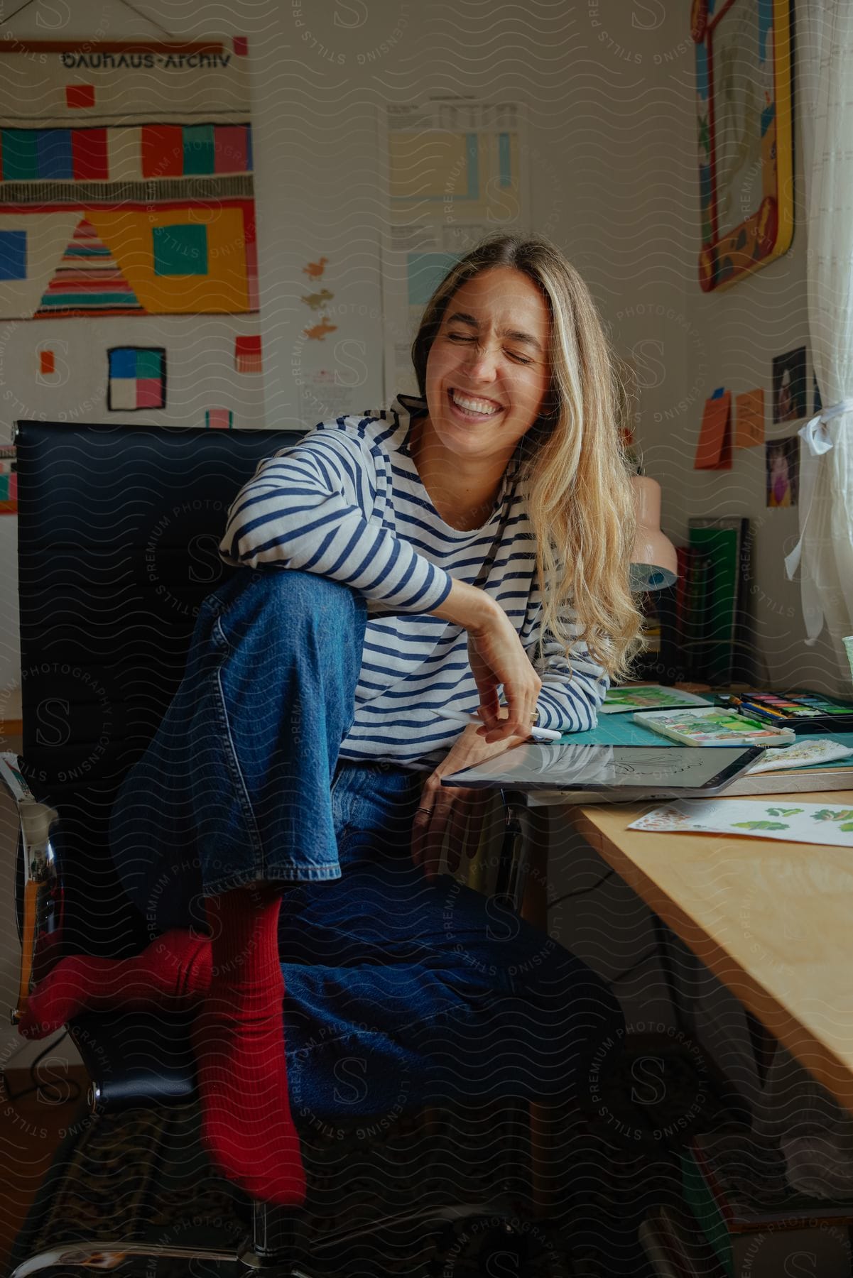 A woman smiles while sitting in a chair by the work table in the art room with a tablet next to her.