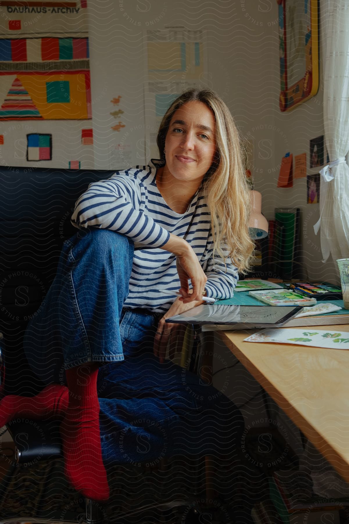 A blonde woman in a striped shirt leans on a worktable with paints on it.