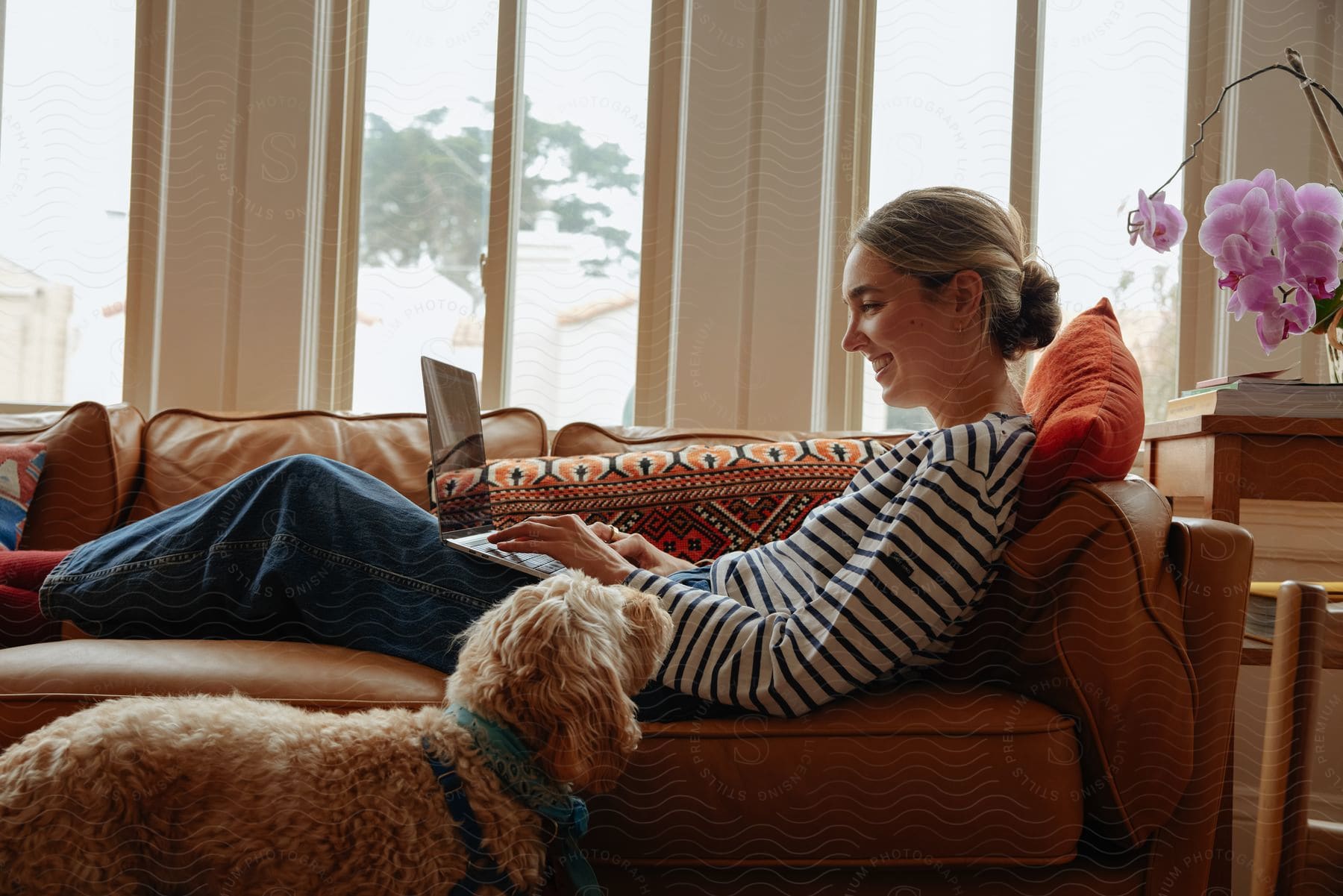 a woman lying on her couch with a laptop on her lap as she smiles while operating it with her dog standing on the floor staring at her