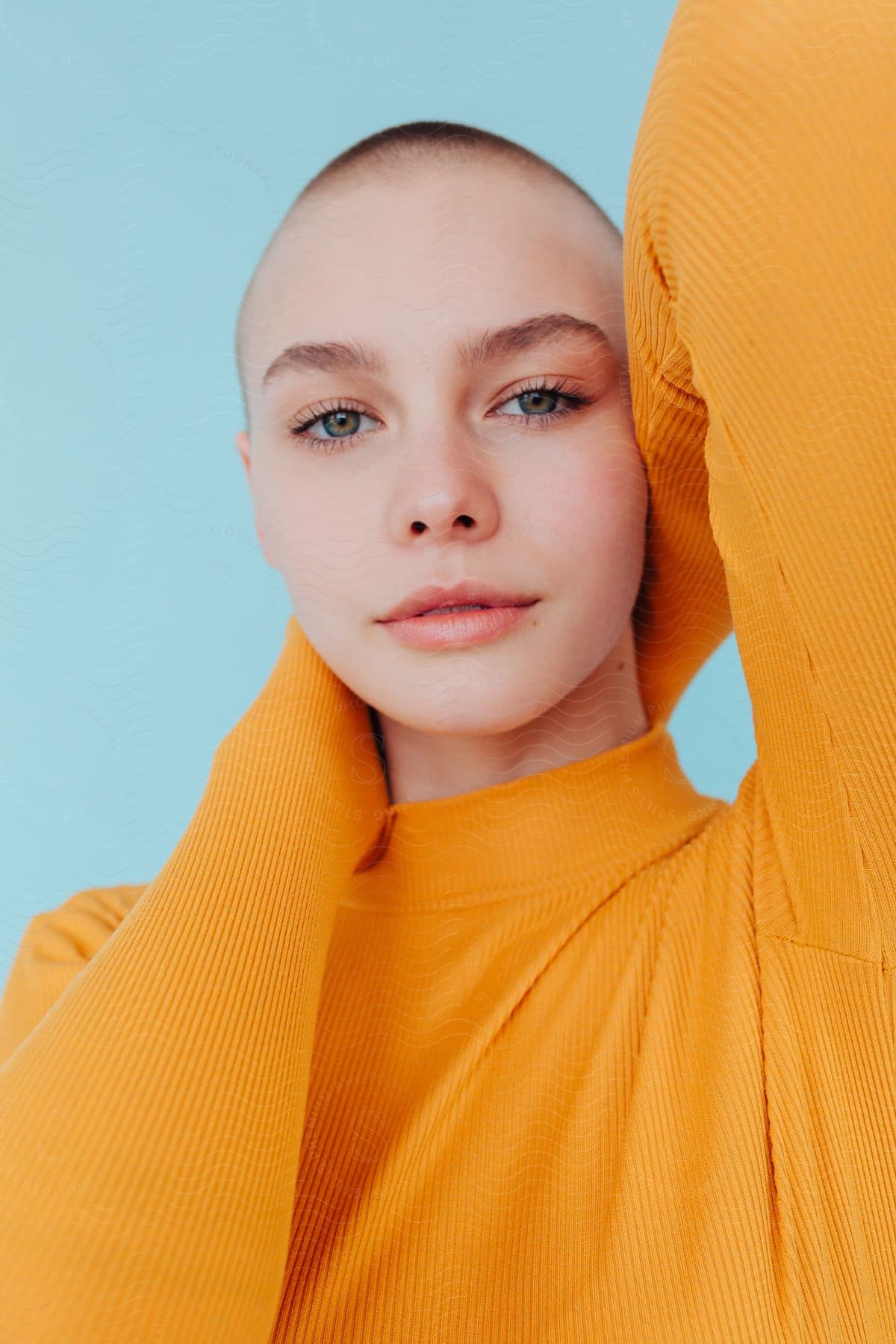 A confident woman with a shaved head and a yellow sweater, posing for a photo with her hands behind her head.