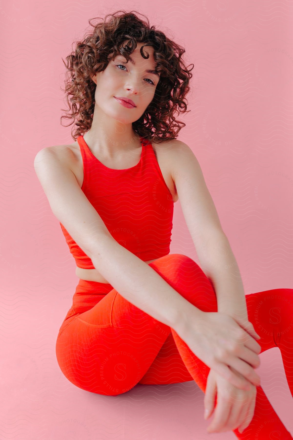 A woman in a red outfit is sitting posing in front of a pink background.