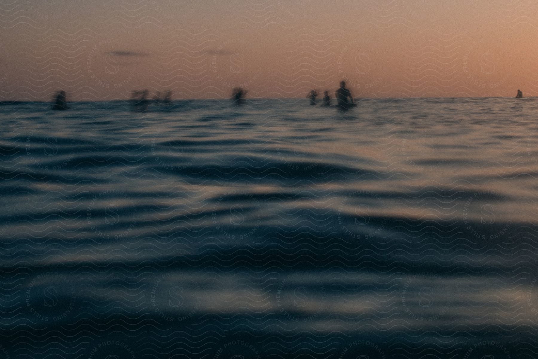 Silhouettes of people in the water and a sailboat in the distance under a sunset sky as light reflects on the ripples and waves