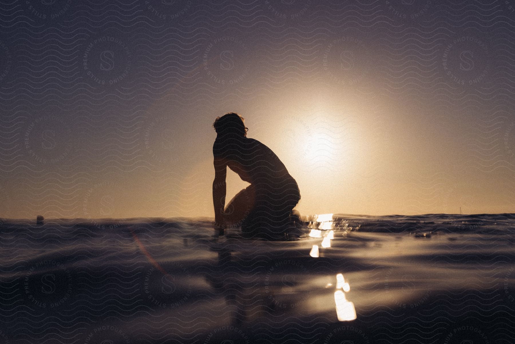 A young man is kneeling on a surfboard in the ocean with the morning sun behind him.