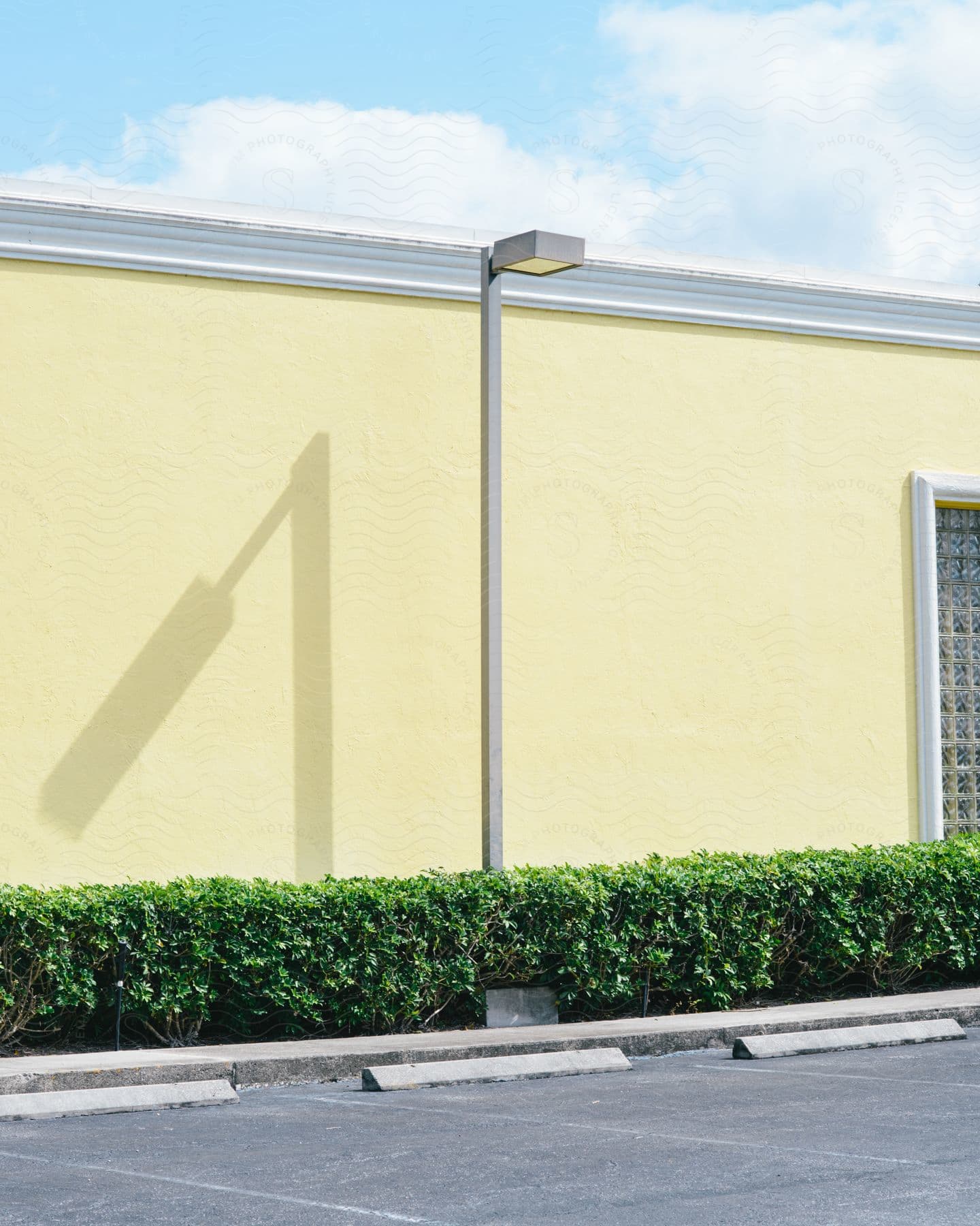 A yellow building with a streetlight shadow on its wall, a parking lot in front, and a green line of small bushes lining the curb.