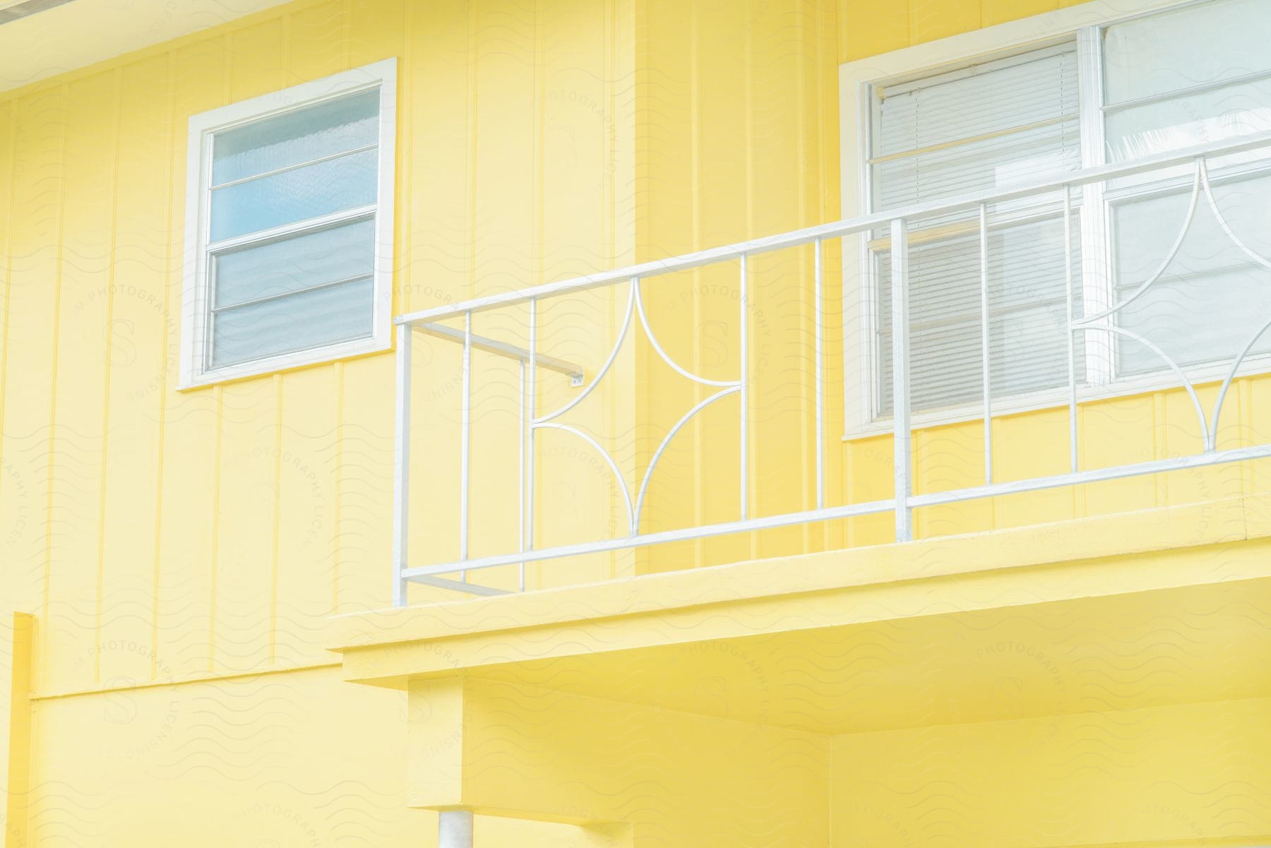 A close-up of a yellow house with a white balcony and windows on the second floor, the windows have white blinds.