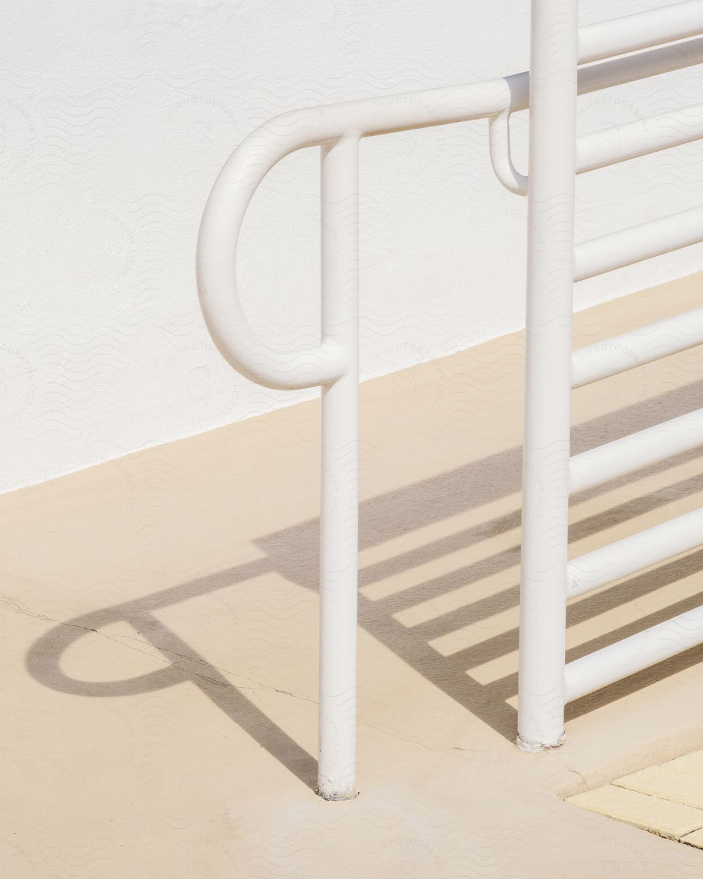 Shadows of a white railing are cast on a cement ramp, between a white wall and the railing.