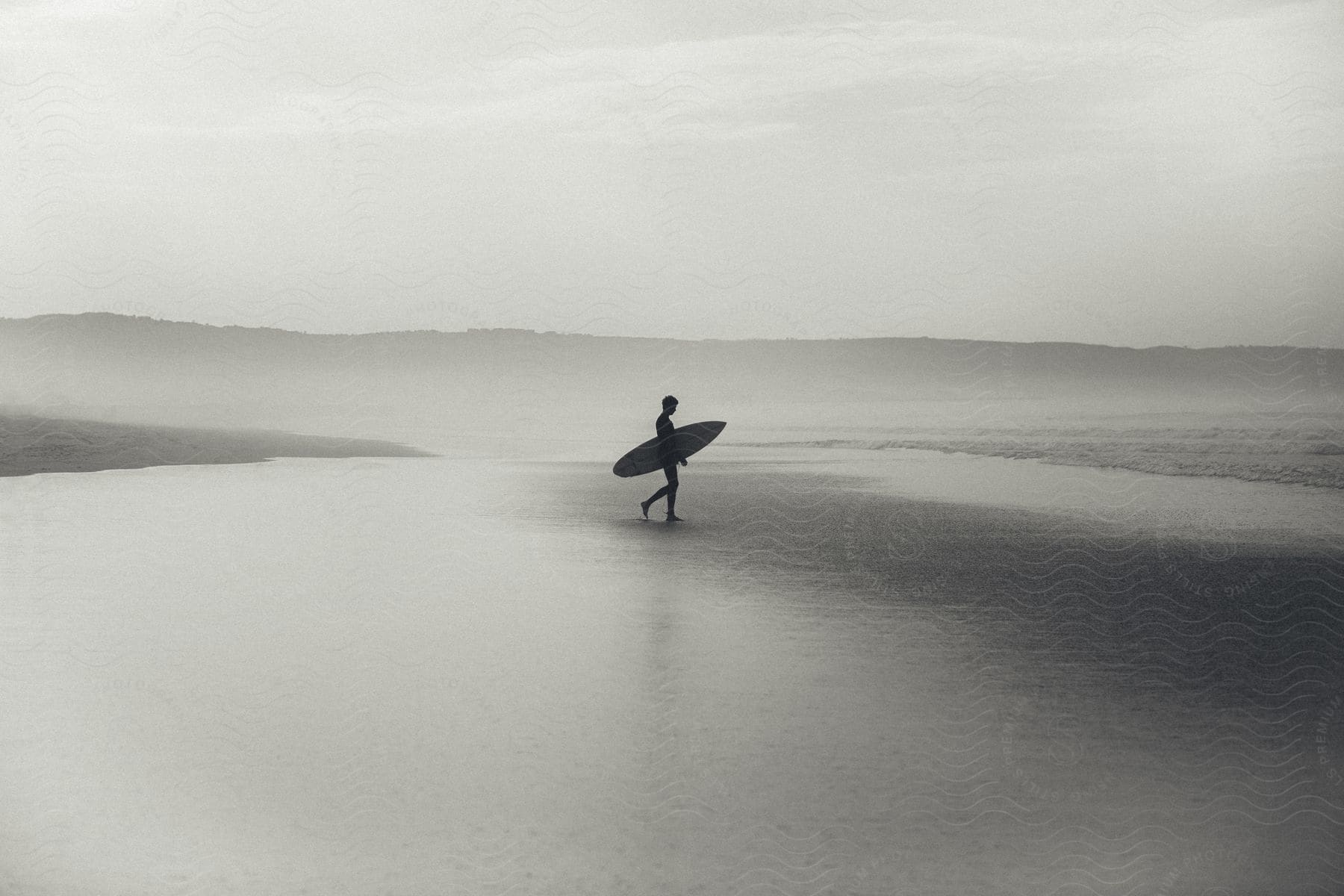 A man is carrying a surfboard as he walks toward the ocean on an empty beach with a mountain ridge in the background on a cloudy day.