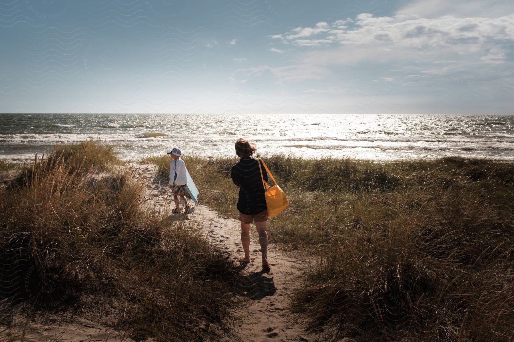 A mother and son walk down a sandy path towards the ocean on a sunny day, carrying a surfboard and a yellow shoulder bag