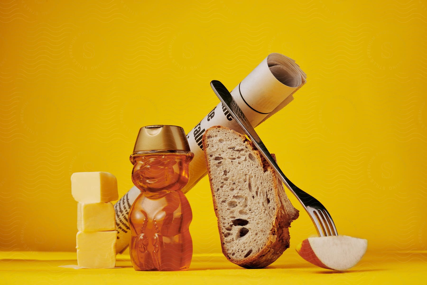 Butter cubes, a bottle of honey shaped like a bear, and a newspaper balanced on a slice of bread kept up by a fork stuck on an apple slice.