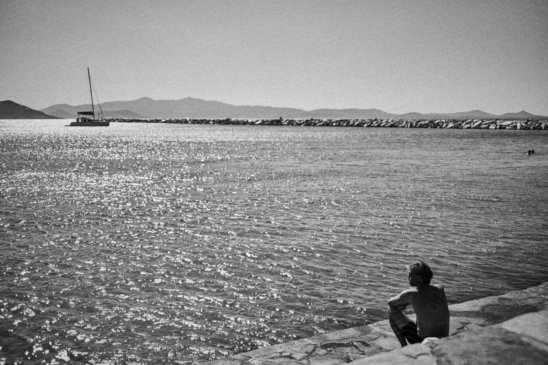 A shirtless man sits on a stone ledge by the water, gazing at a distant boat with mountains in the background and a rocky jetty to the right.