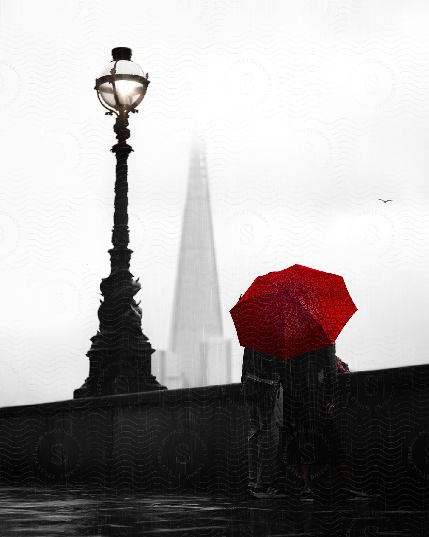 A couple on a bridge next to an old lamppost shelter under a red umbrella while looking out at a skyscraper obscured by clouds.