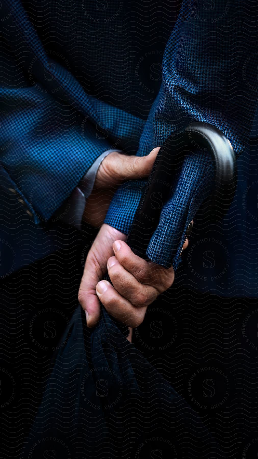 A man holds the handle of an umbrella in his hands behind his back while he's wearing a stylish blue suit.