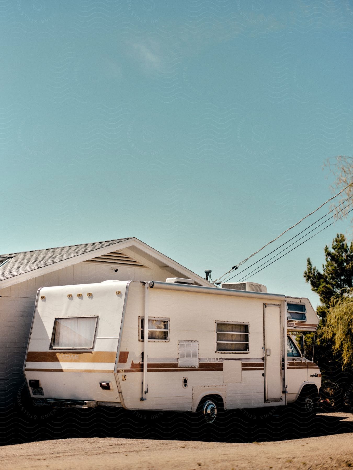 A vintage 1970s white camper, parked by a house on a dirt road on a sunny day.