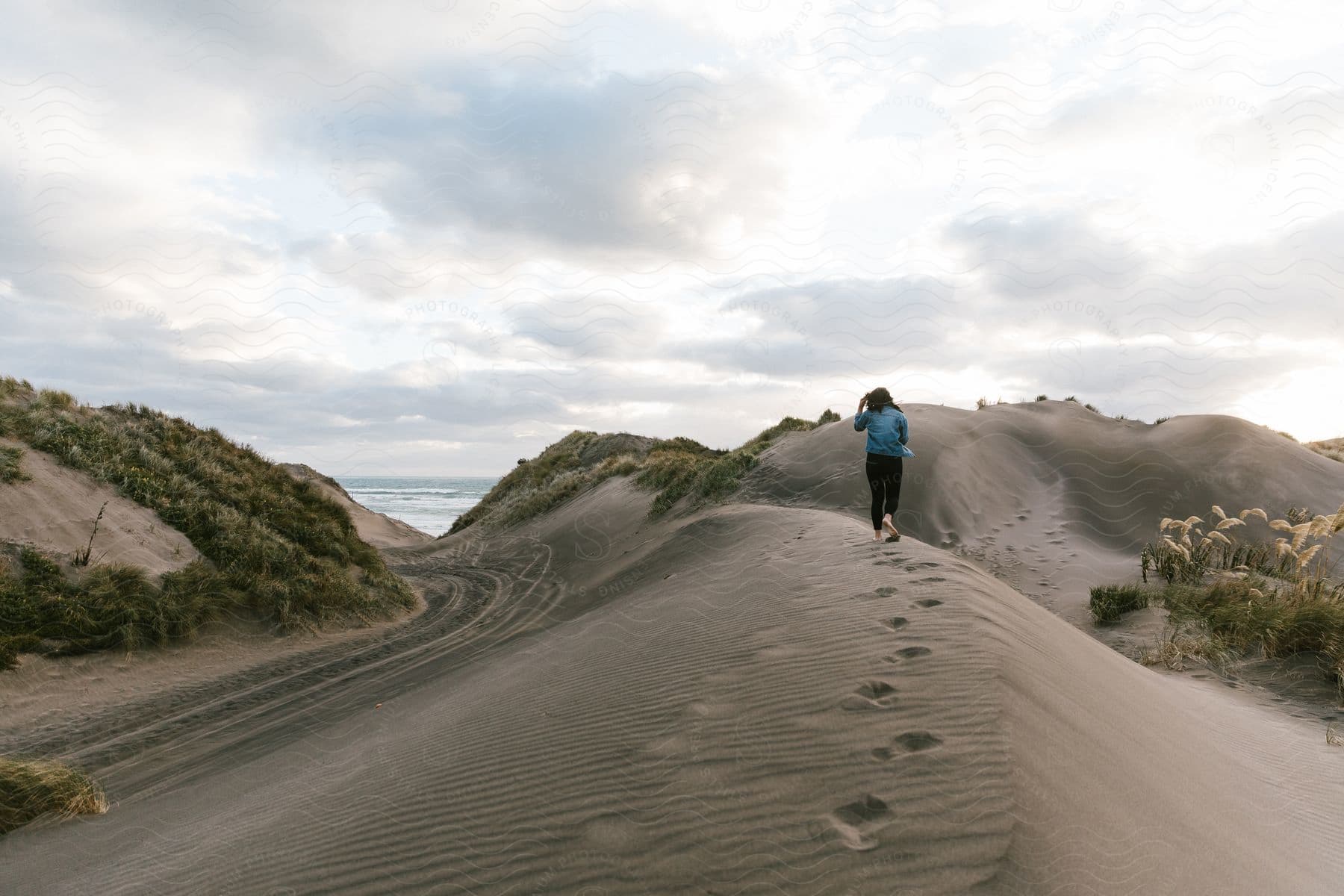 A woman walking barefoot over a sand dune towards a beach.