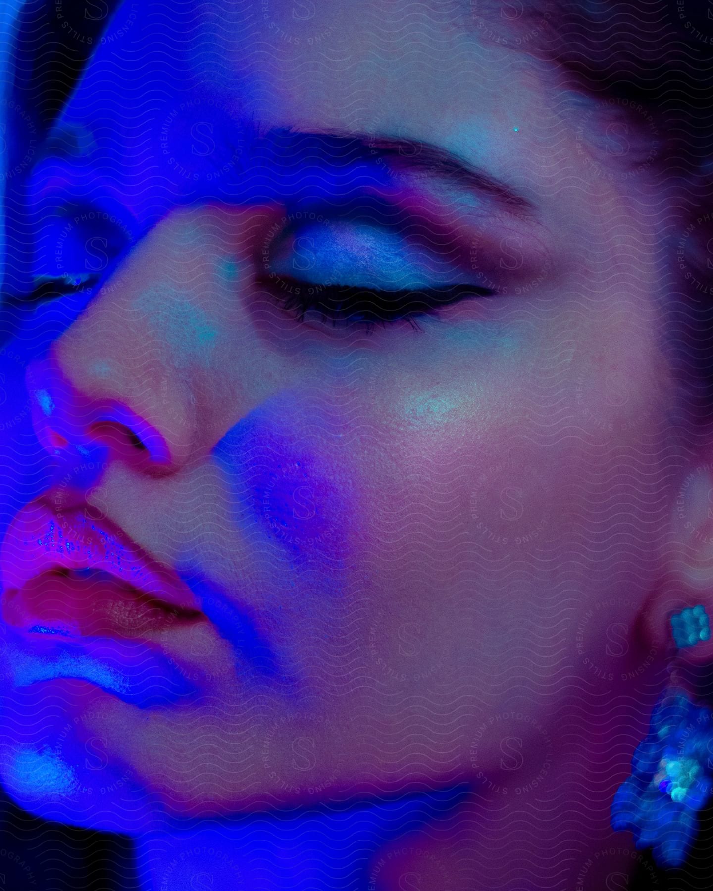 In a softly illuminated room, a young woman's face is adorned with makeup. The bluish light accentuates her features, and she wears elegant blue designer earrings