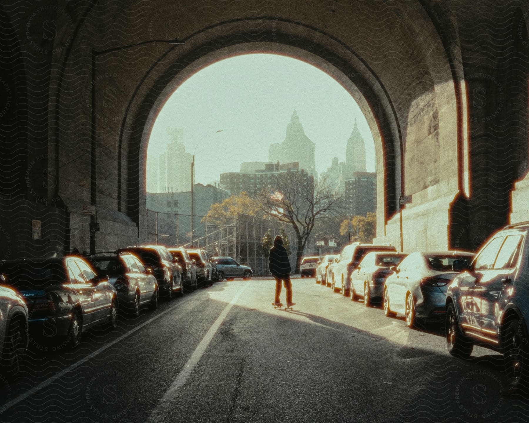 Person in a hoodie on top of a skateboard inside a tunnel with parked cars on the sides and a city with tall buildings in the background.
