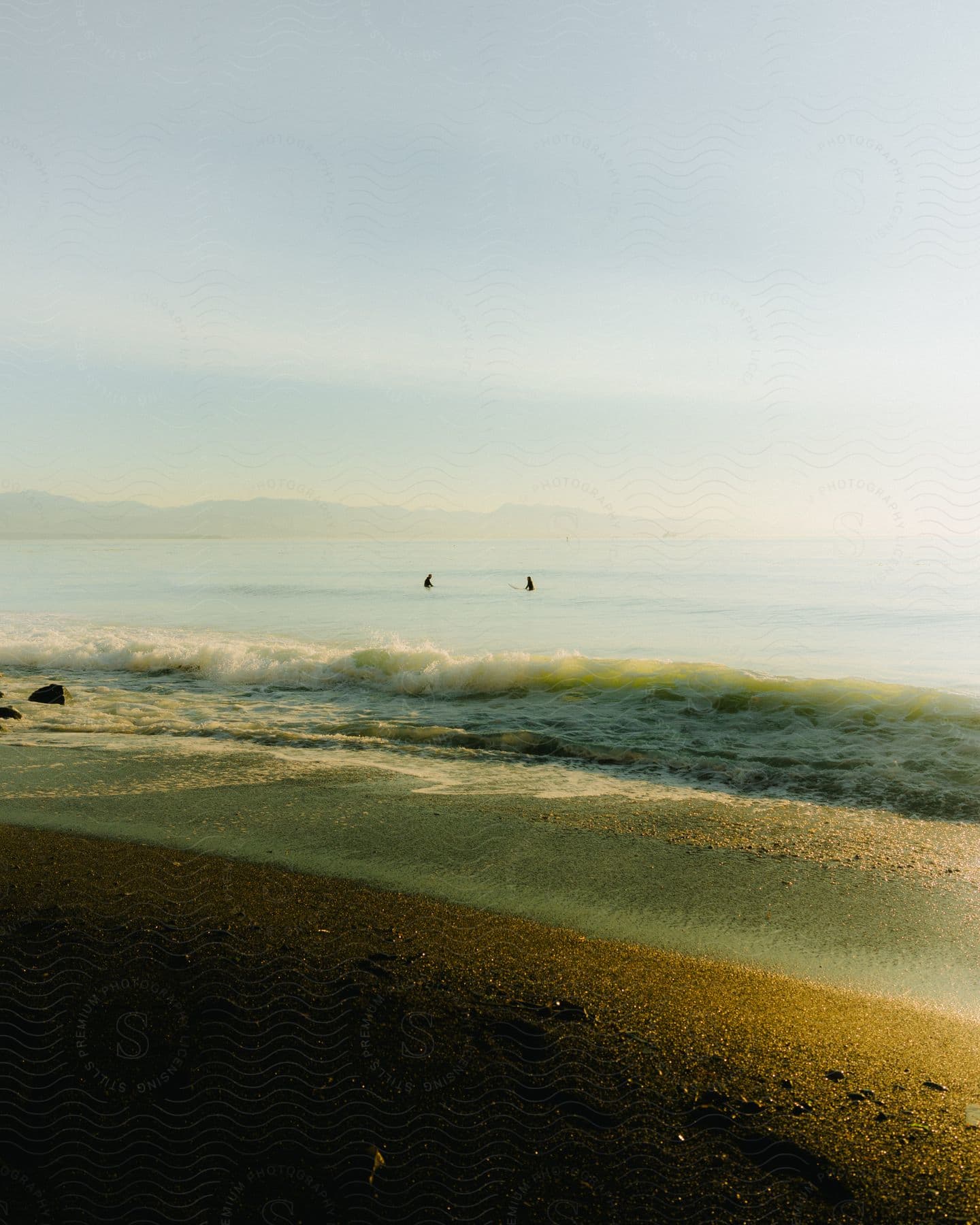 Waves roll into shore as two people are out in the water under a hazy sky