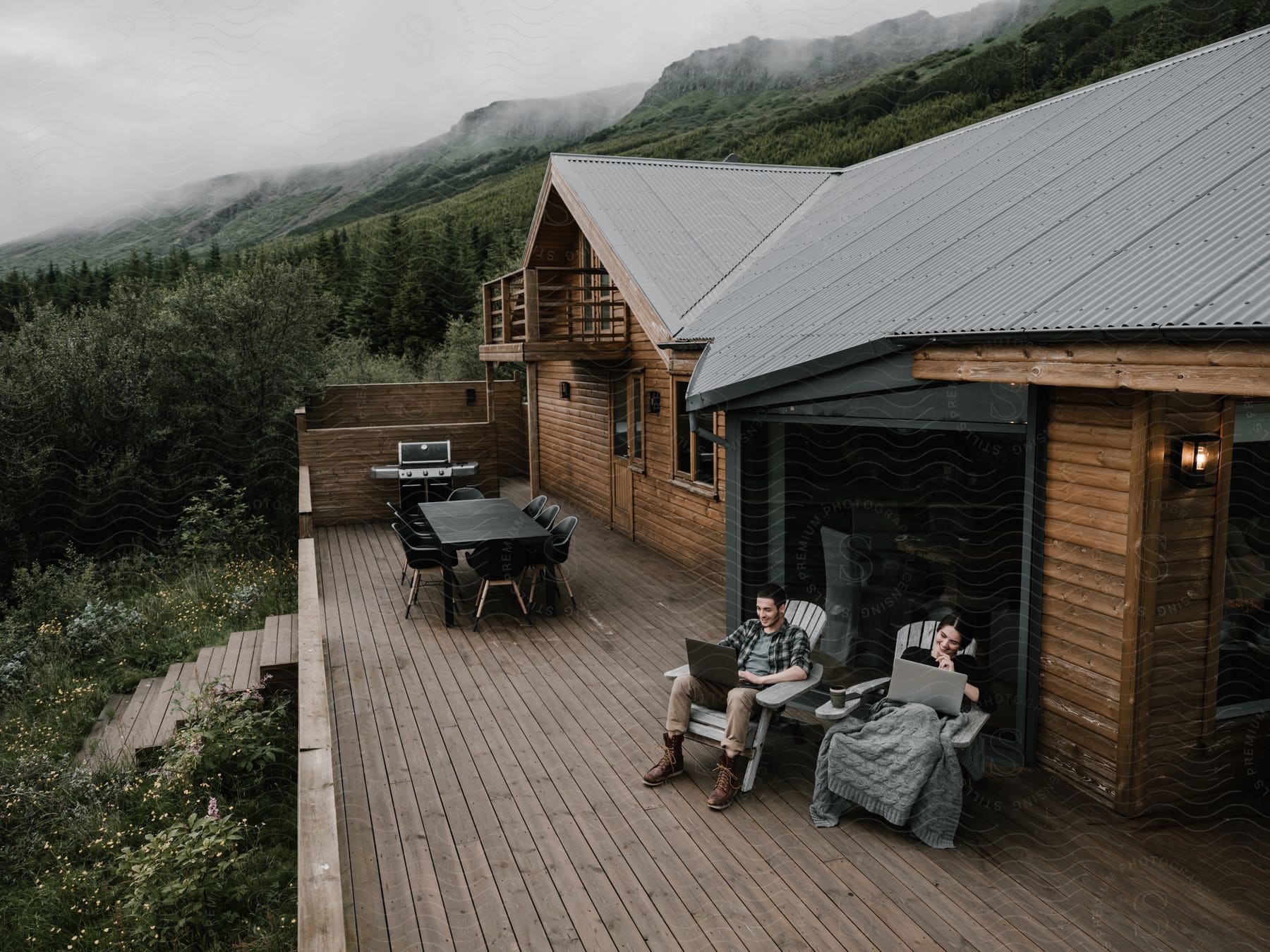 A couple works on their laptops on a deck overlooking a mountain range shrouded in mist and fog, with a grill and a table nearby.