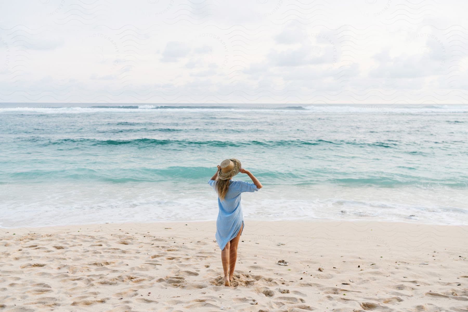 The backside of a woman standing on a sandy beach holding her sun hat as she looks out at the ocean waves