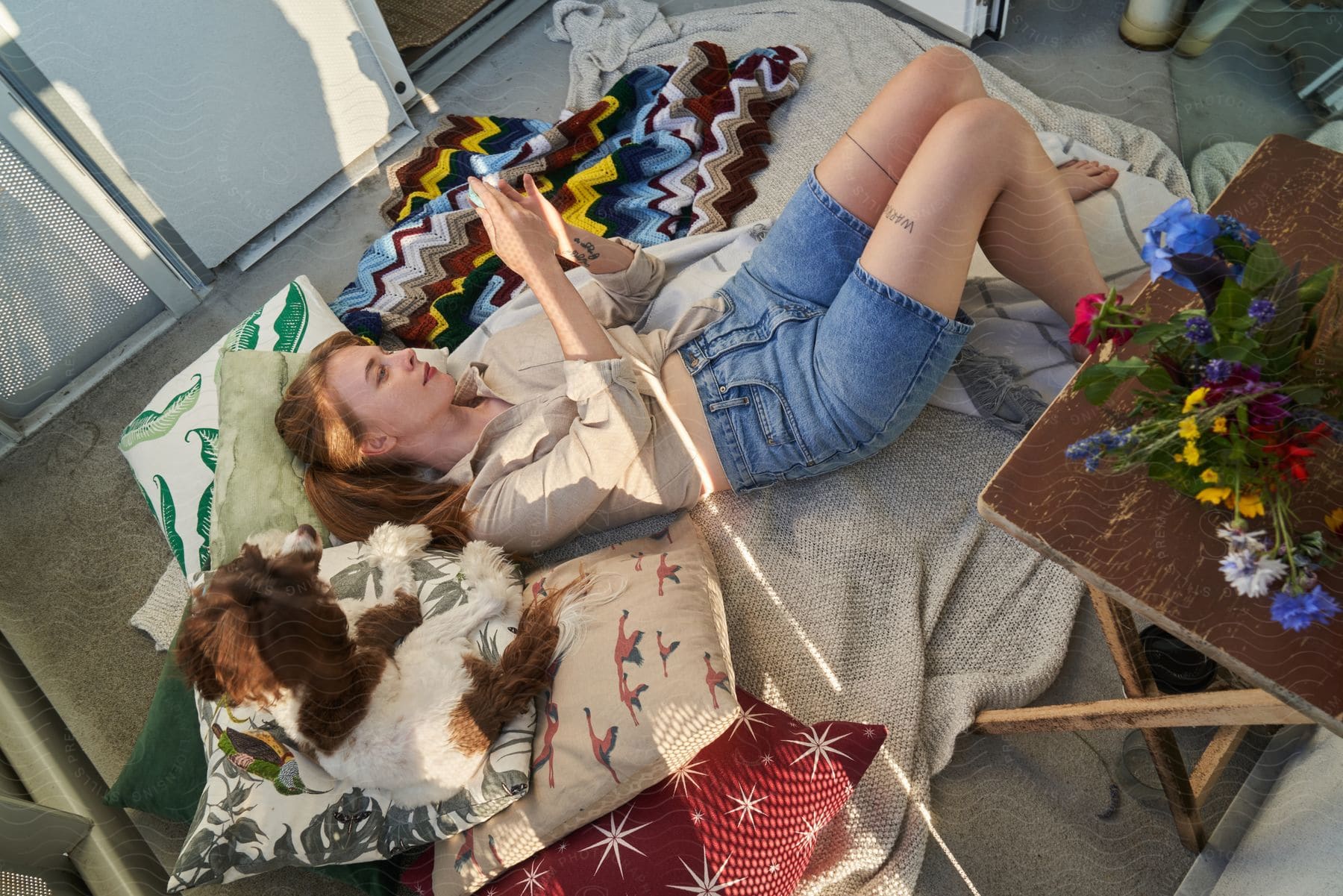 A smiling young woman lying on a blanket on the floor as a dog watches her.