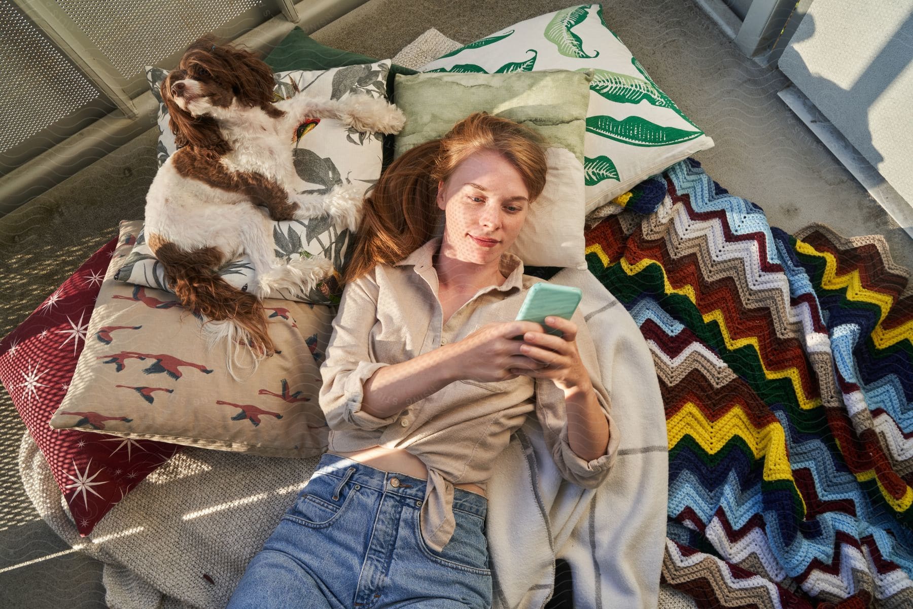 A woman laying on her bed against pillows as she looks at her cell phone with her dog laying next to her