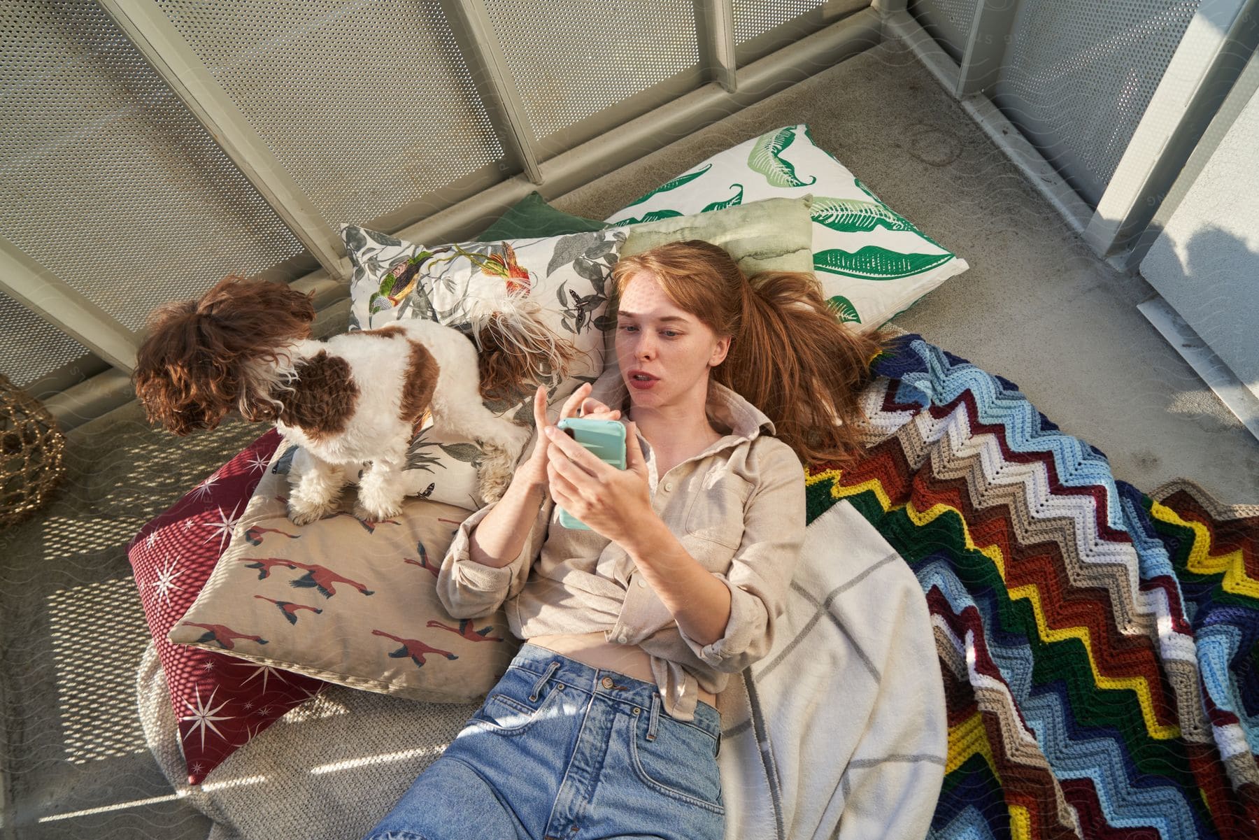 A young woman next to her dog is checking her phone while laying down on a blanket on the balcony.