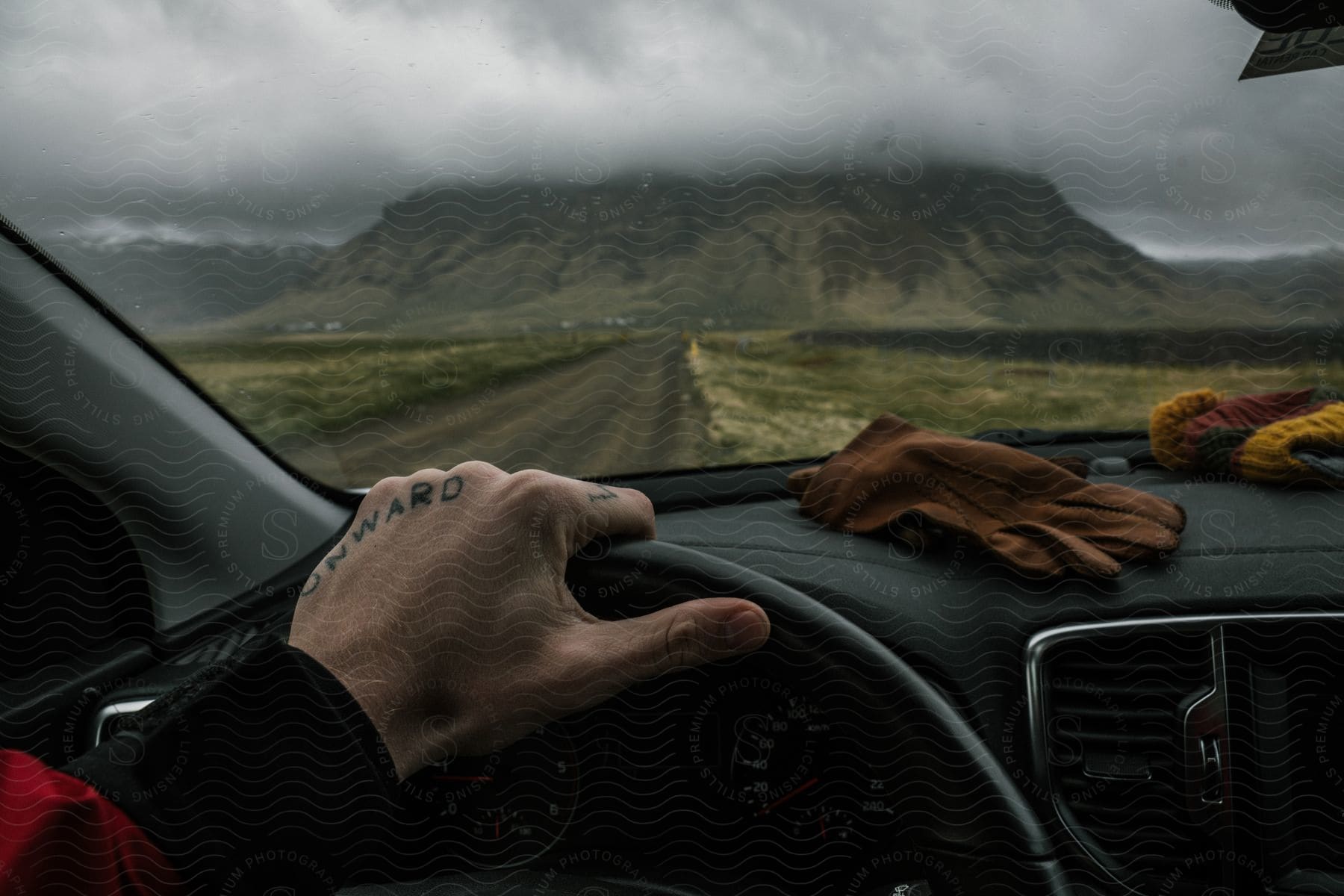 Close-up of a hand on the car's steering wheel, with a mountain covered by clouds ahead.