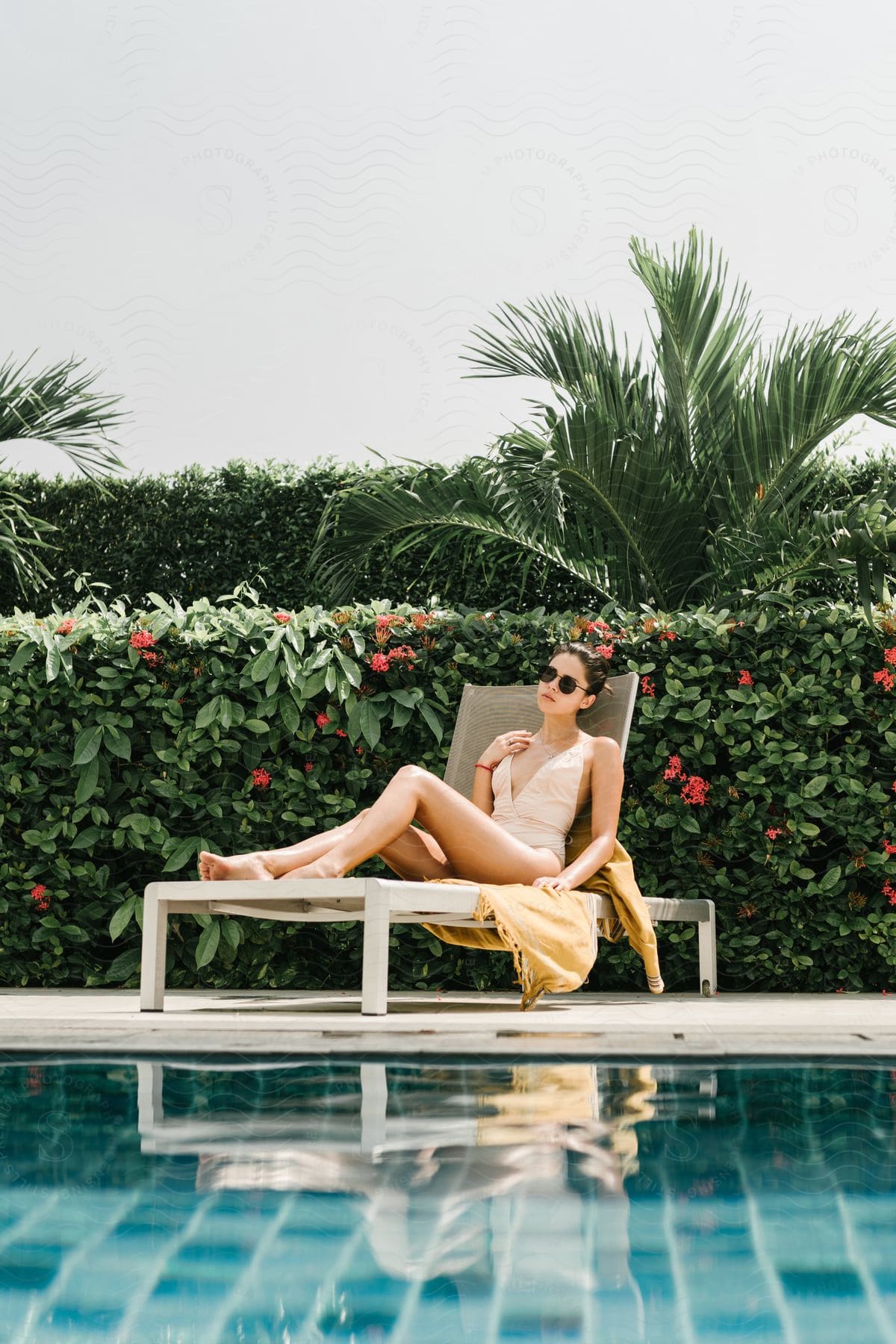 Stylish young woman outdoors on a bathing suit wearing sun glasses sitting next to a pool surrounded by plants and flowers during day time