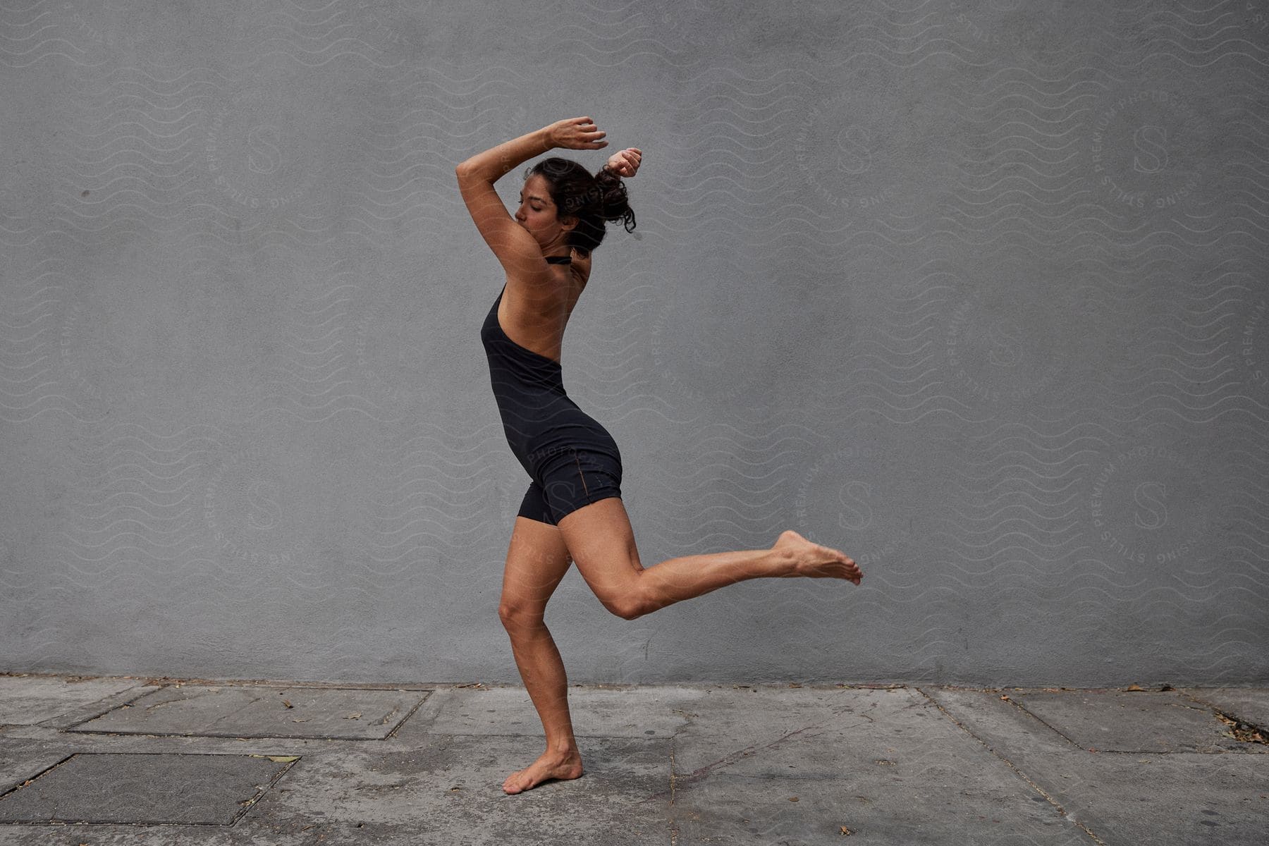 Woman with a black top and shorts dances in front of a gray wall on the street