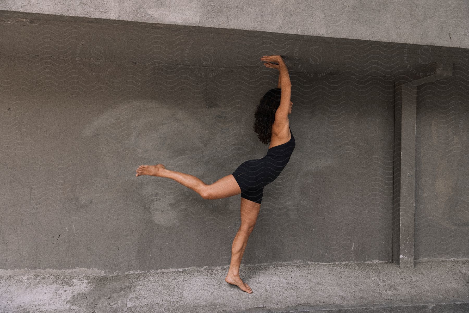 a woman in a black jumpsuit poses with one leg outstretched and her arms against the ceiling.