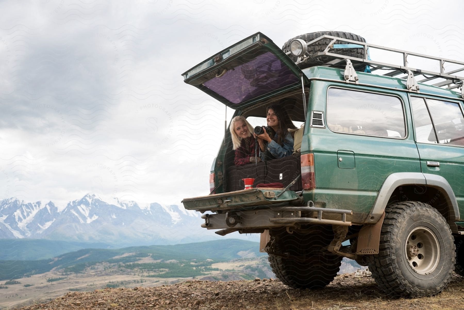 Two women are sitting in the back of a jeep smiling as one woman holds a camera with mountains in the distance