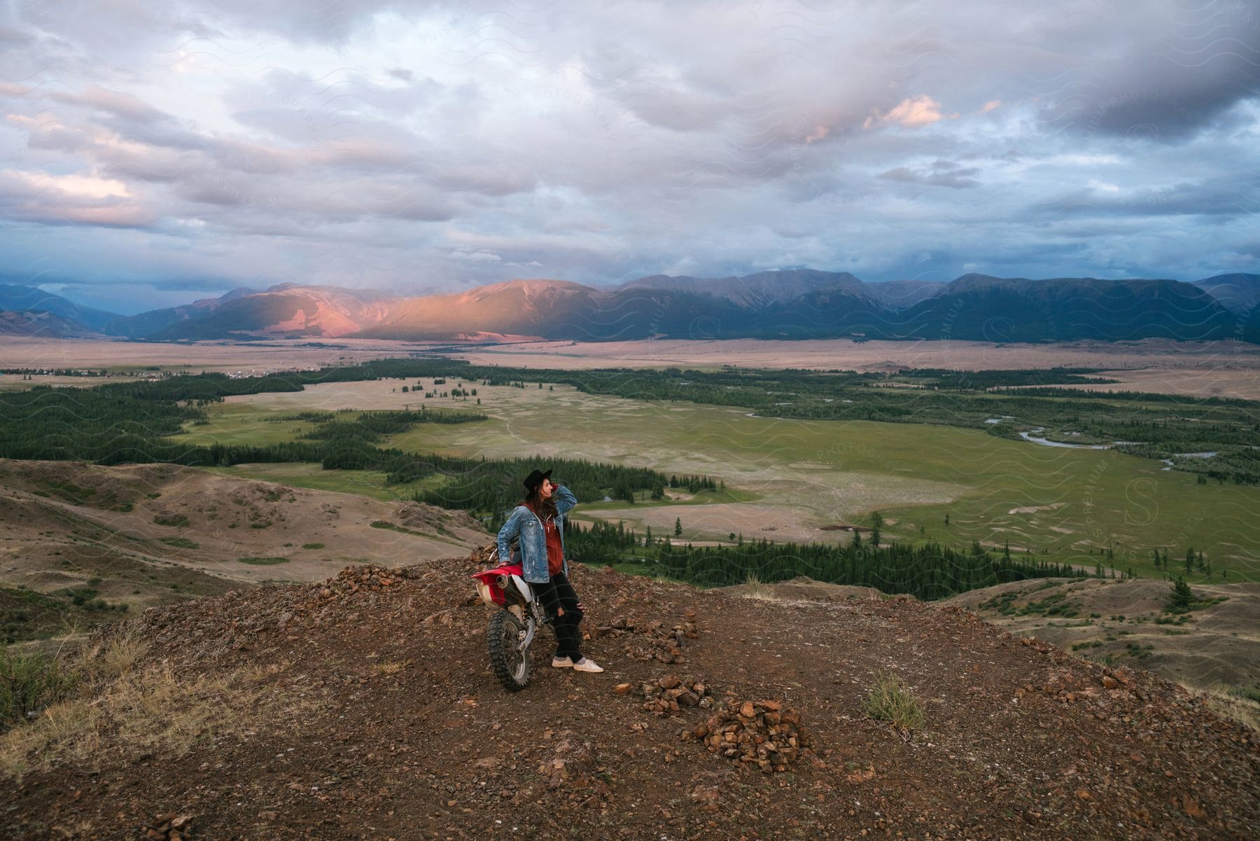 A teenage girl is leaning against her dirt bike on a hilltop with mountains in the distance