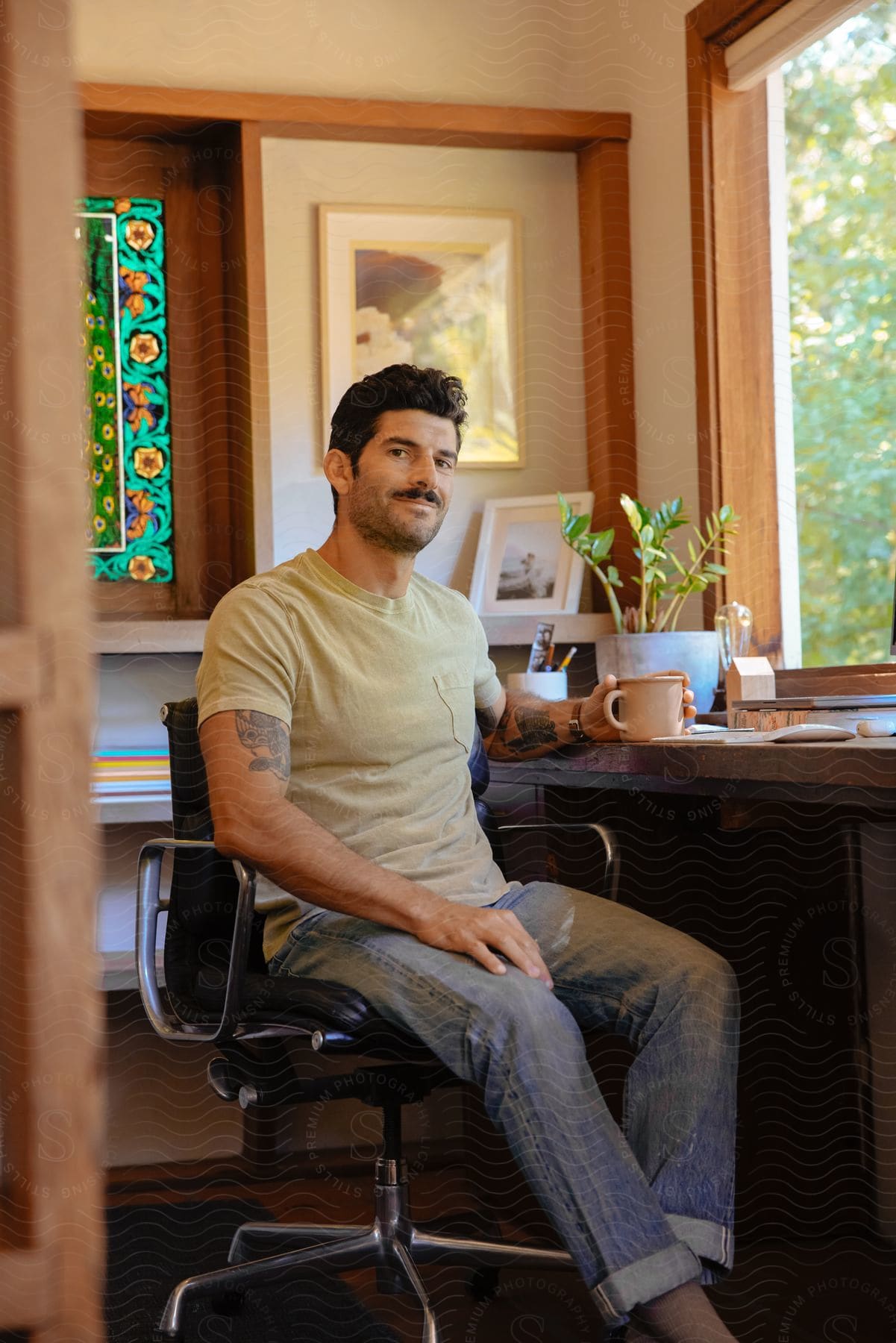 Stock photo of a man in jeans and a shirt sits at his home office desk, facing the camera, with a closed laptop and a cup of coffee in hand.